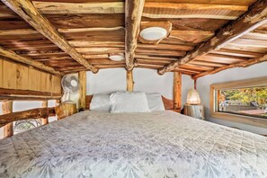 A rustic wooden cabin bedroom with a bed, a fan, and a window with a view.