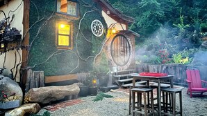 Ein rustikales Haus mit einem Rundfenster, einer Tür und einer kleinen Sitzgelegenheit im Freien mit Tisch und Stühlen.