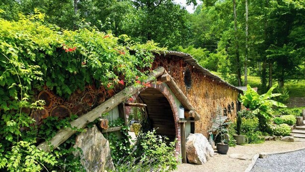 Ein rustikales Steinhaus mit einem hölzernen Rundbogenportal, umgeben von üppiger Vegetation und einem Kiesweg.
