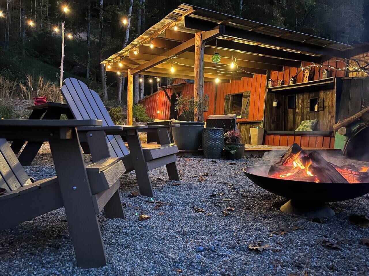 Una terraza de madera con sillas Adirondack, un hogar para fuego y luces colgantes.