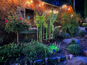Surrounded by lush greenery, this charming outdoor space features a stone wall, a wooden door, and a quaint wooden structure with twinkling string lights.