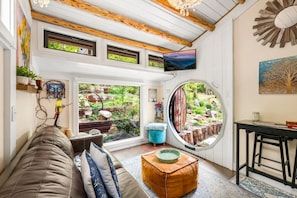 Ein gemütliches Zimmer mit einer Couch, einem runden Tisch, einem Fernseher und einem großen Fenster mit Gartenblick.