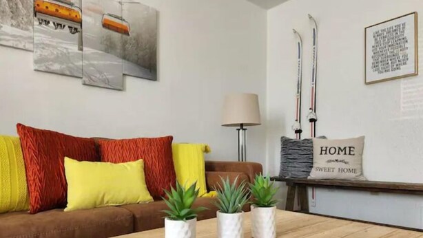 Ein Wohnzimmer mit braunem Sofa, gelben und roten Kissen, drei Topfpflanzen, einem Couchtisch aus Holz mit Fernbedienung sowie Wandbildern, darunter ein gerahmtes Zitat und ein Paar Ski.
