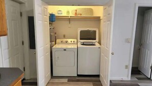 Ein Waschraum mit Waschmaschine und Trockner, eine kleine Ablage mit Reinigungsmitteln und eine Tür zu einem weiteren Raum.