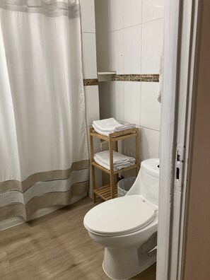 Ein Badezimmer mit Toilette, einem hölzernen Ablagebrett mit gefalteten Handtüchern, einer Duschvorhang und gefliesten Wänden.