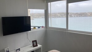Quarto com televisão na parede, uma pequena estante branca com moldura de foto e uma janela com vista para uma vila costeira e o mar.