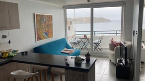 Cozinha moderna com sofá azul, área de jantar com cadeiras e varanda com vista para o mar.