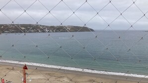 Vista costeira através de uma vedação de arame, mostrando uma praia, mar calmo e colinas ao longe.