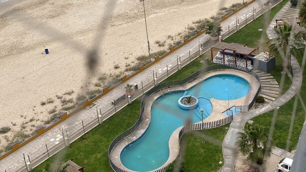 Uma piscina com design curvo singular, uma praia com pessoas e um horizonte urbano ao fundo.