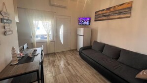 A living room with a dark grey sofa, a wooden dining table with chairs, a flat-screen TV mounted on the wall, and a window with sheer curtains. - Vacation apartment, quiet part. Perfect for your vacation! (Vir)