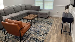 A modern living room with a grey sectional sofa, a brown leather chair, and a small ottoman. There is a blue patterned rug on the wooden floor. - Spacious 4-bedroom house  walking distance to Mercedes Benz Stadium & Downtown (Atlanta)