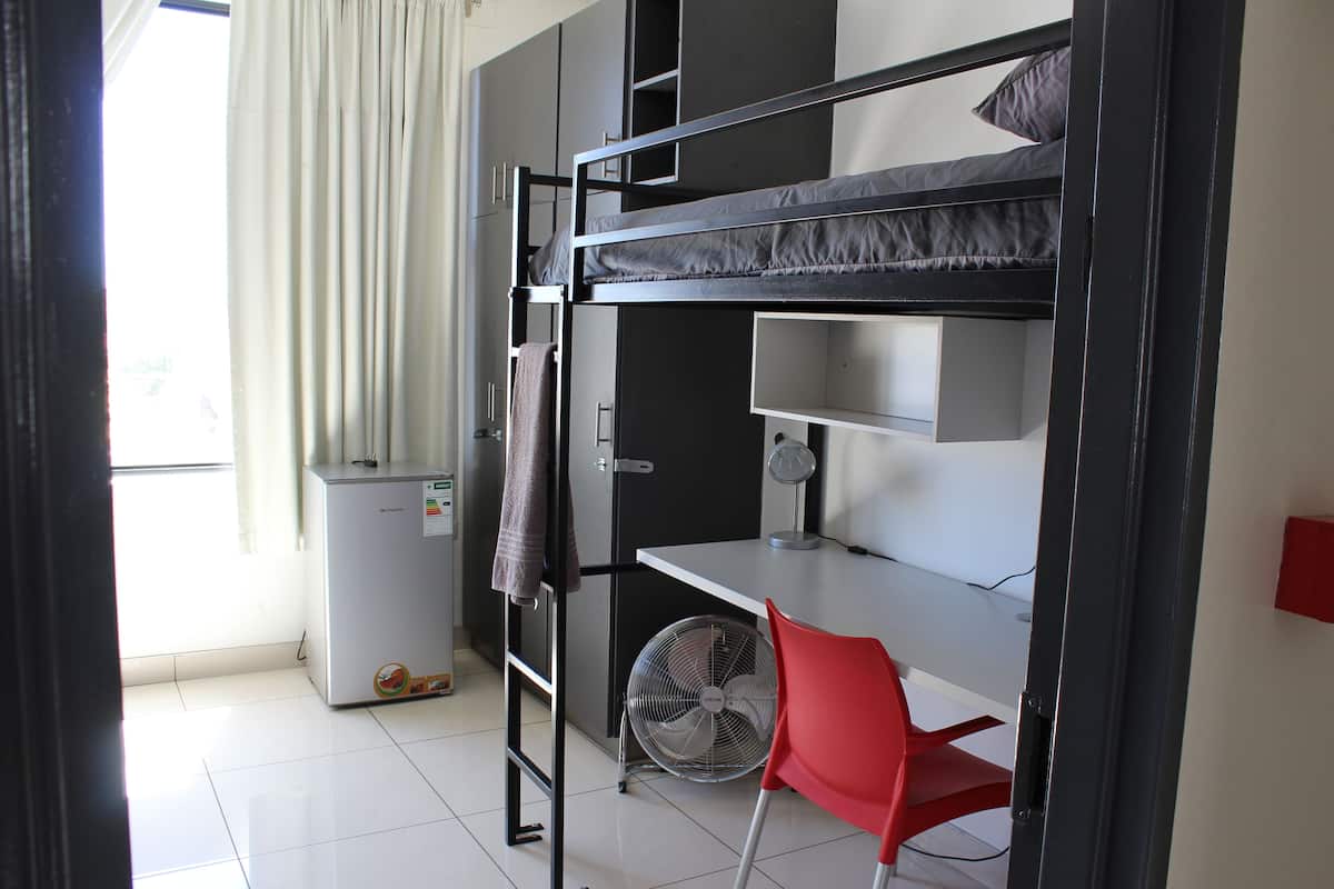 A dormitory room with bunk beds, a desk, a chair, a fan, and a small refrigerator.
