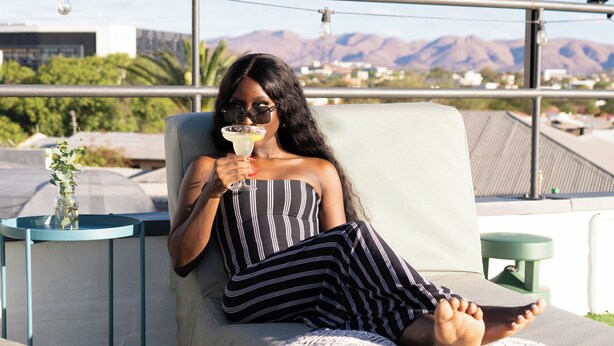 A person relaxing on a lounge chair with a drink, overlooking a cityscape with mountains in the background. - TaTe Village - Hostel (Windhoek)