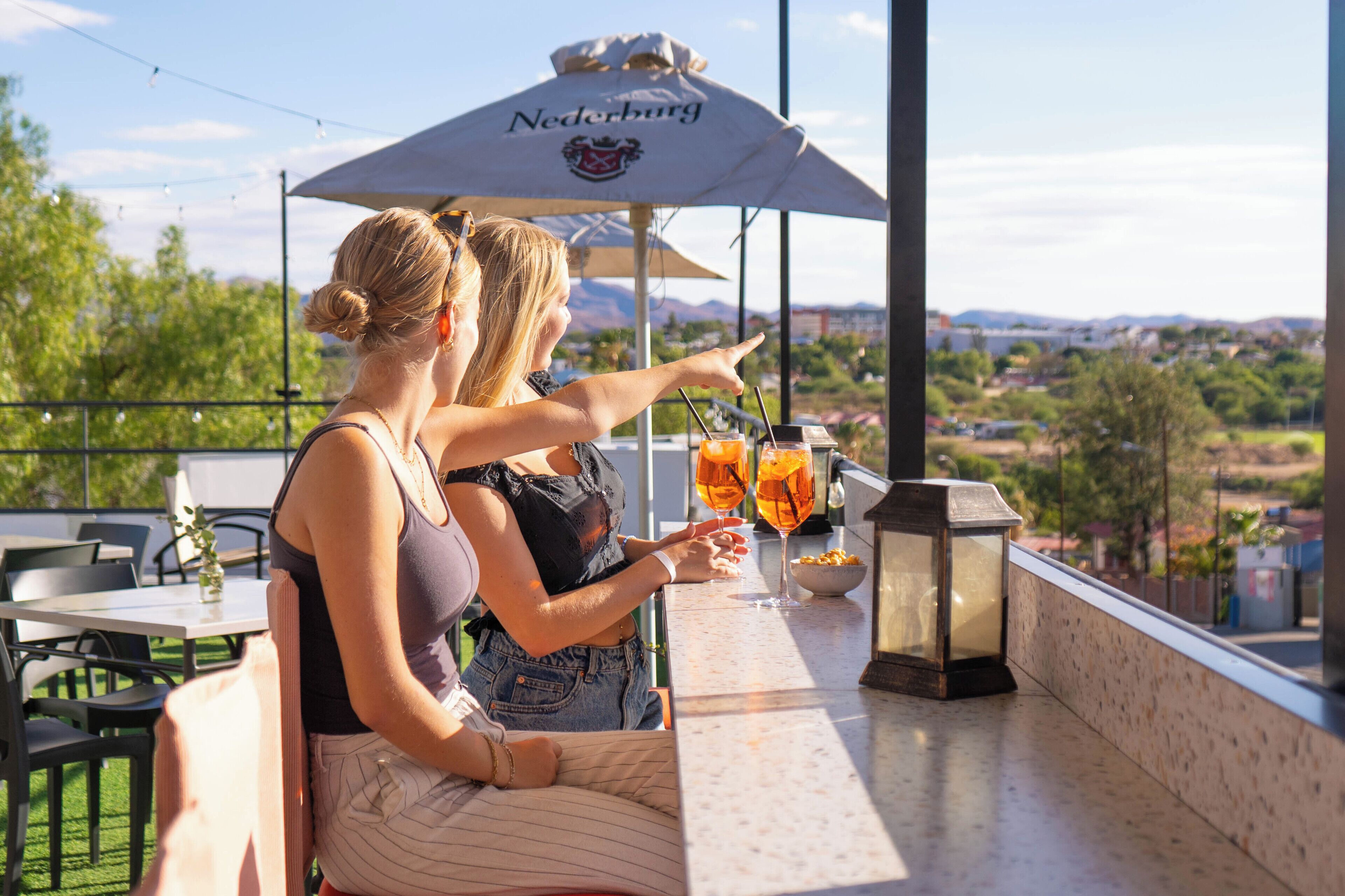 Deux femmes sont assises à un bar en plein air, d’où l’on a une vue sur la ville.