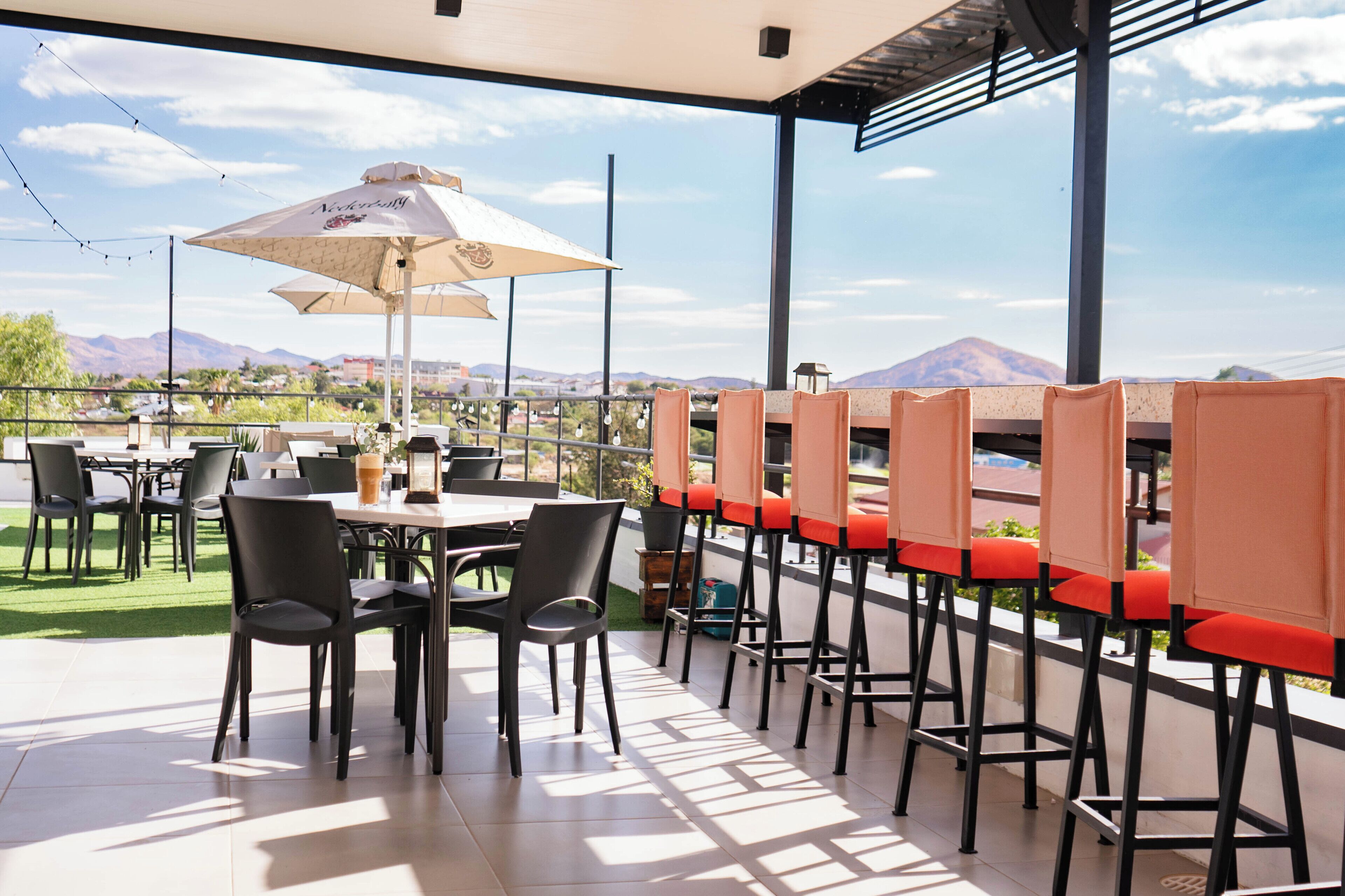 An outdoor dining area with tables and chairs, a large umbrella, and a view of mountains.