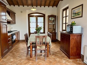 A kitchen with wooden cabinets, a dining table with chairs, a microwave, and a window with a view of greenery. - Villa with private pool and panoramic view in beautiful Val d'Orcia (San Giovanni d'Asso)