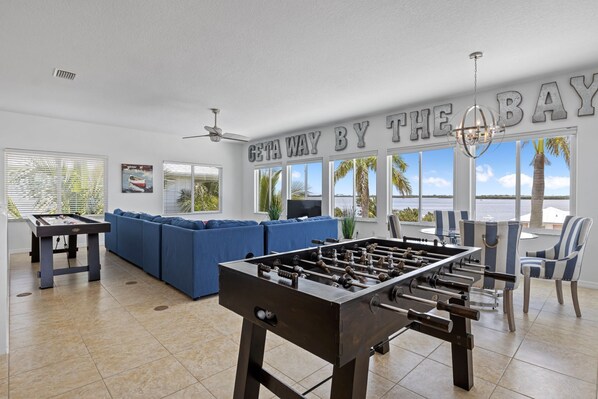A spacious living room with a foosball table, a blue sofa set, and a large window offering a view of the water.