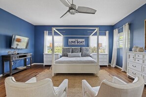 A bedroom with a four-poster bed, a ceiling fan, a TV, and a window with blinds.