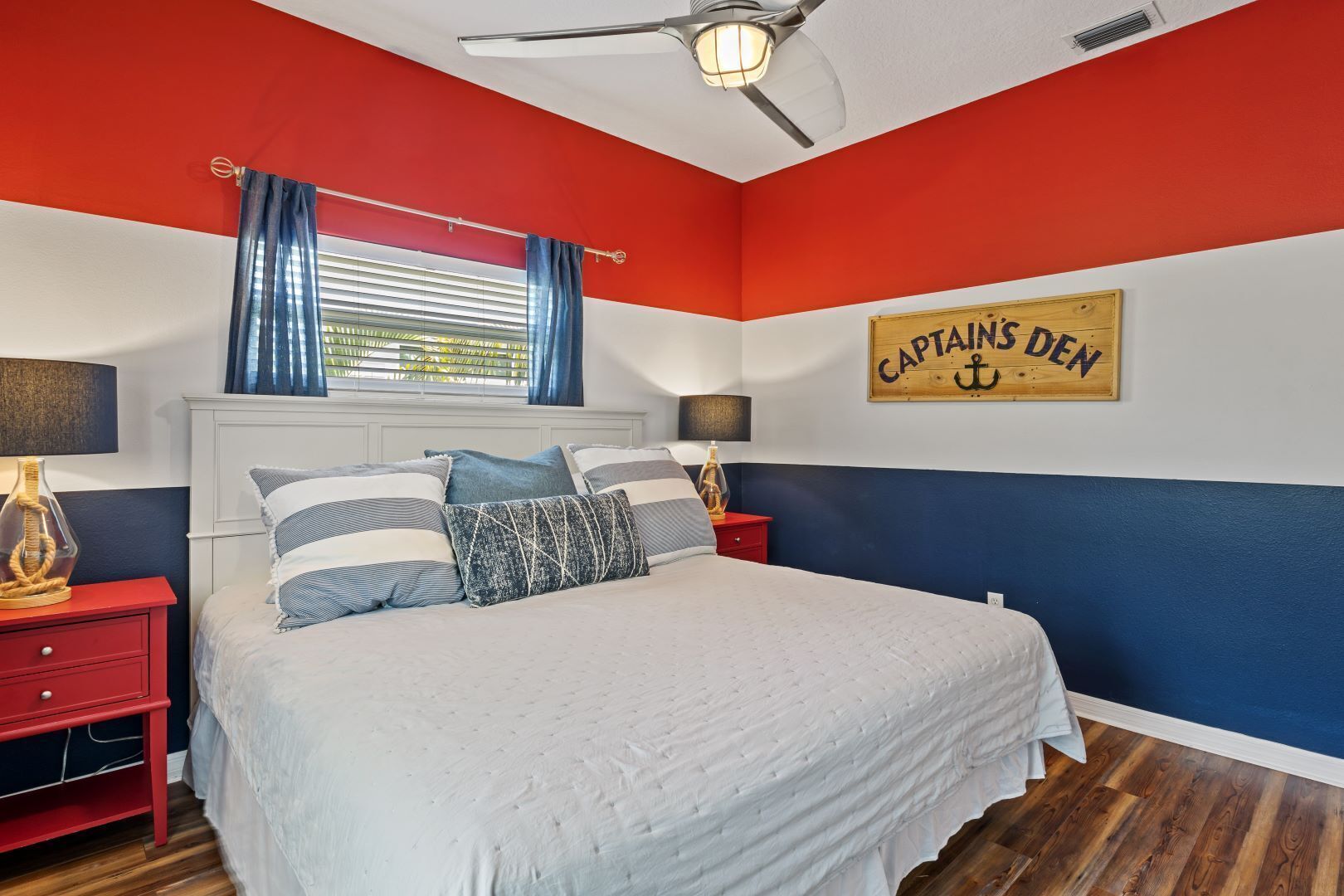 A bedroom with a bed, red and white walls, a ceiling fan, a window with blue curtains, and a sign that reads "CAPTAINS DEN".