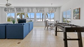 A spacious living area with a large sofa, a foosball table, and a dining table with striped chairs. A large window offers a view of the ocean. - Getaway By The Bay-4 bedrooms-Large private pool and spa across from the bay!! (Bradenton Beach)