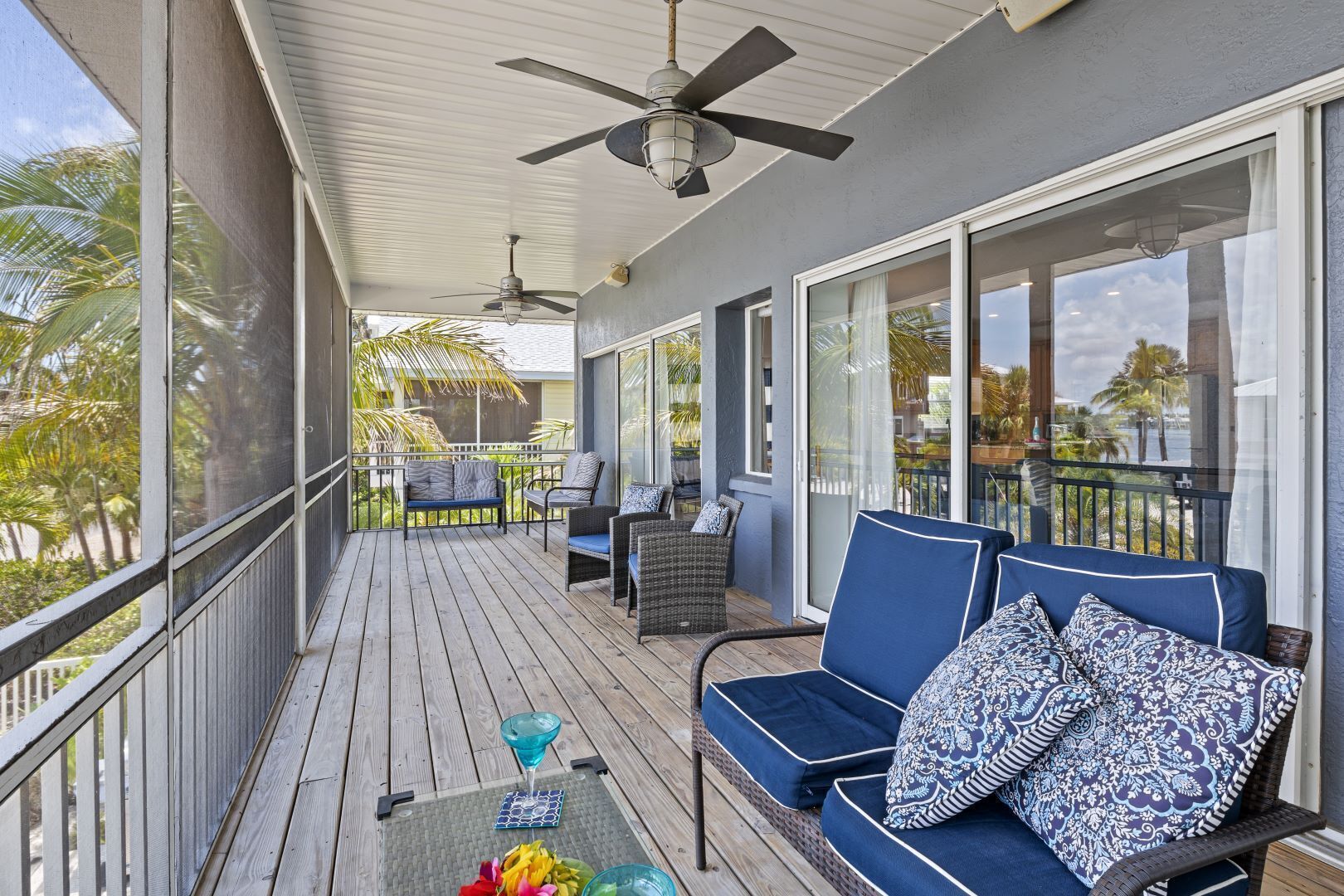 A covered outdoor balcony with a ceiling fan, a seating area with blue cushions, and a glass table with a drink and fruit.