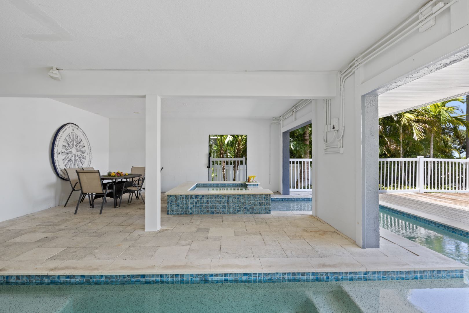 A covered outdoor swimming pool area with a hot tub, a dining table with chairs, and a compass wall decoration.