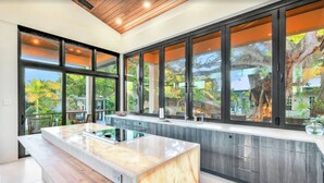 A modern kitchen with a large island, dark cabinetry, and large windows offering a view of the outdoors.
