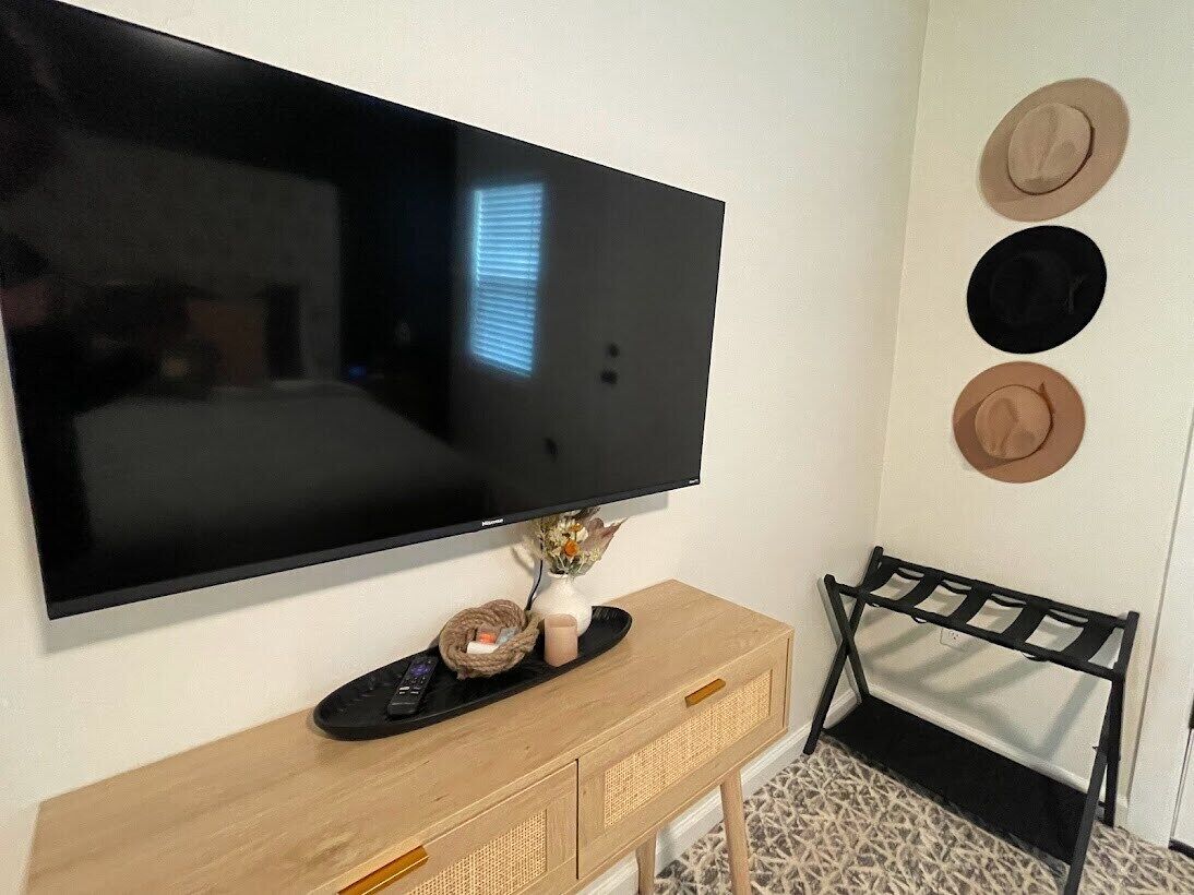 A TV mounted on a wall, a wooden console table with a remote, and three hats on the wall.