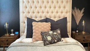 A bedroom with a tufted headboard, a bed with multiple pillows, a nightstand with a lamp, and a wall clock. - Boho 405 - DI Home Rentals (Stillwater)