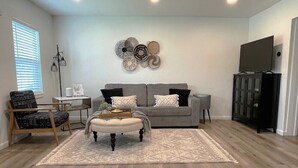 A living room with a grey sofa, a wooden chair, a small table, and a TV on a cabinet. - Boho 405 - DI Home Rentals (Stillwater)