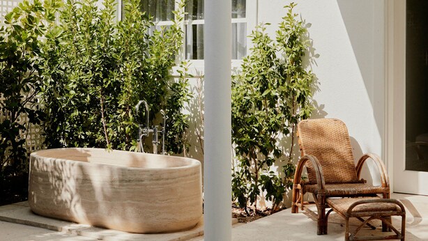 A white building with a large, round outdoor bathtub, a wicker chair, and lush greenery.