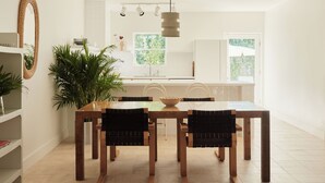A dining area with a wooden table and four chairs, a large potted plant, and a kitchen in the background.