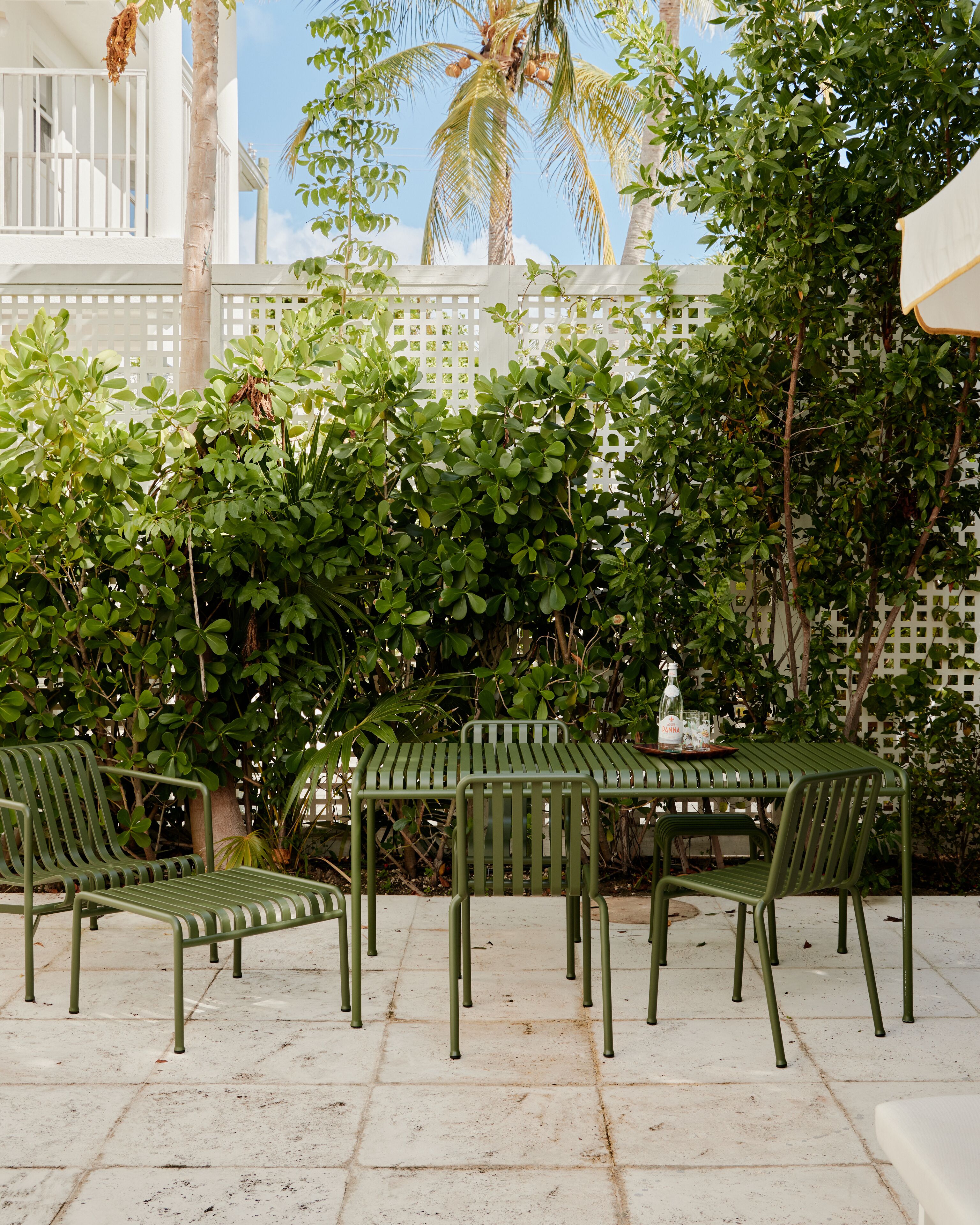 A patio with a green metal table and chairs, surrounded by lush greenery and a white fence.