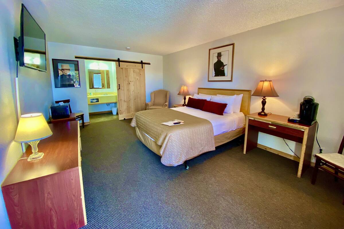 Ein Hotelzimmer mit einem Bett, Nachttischlampen, einem Schreibtisch mit Lampe, einem Sessel, einer Kaffeemaschine, einem Spiegel, einem Bilderrahmen und einer Schiebetür.