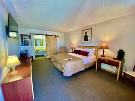Habitación de hotel con cama, lámparas de noche, escritorio con lámpara, una silla, cafetera, espejo, marco de fotos y puerta corrediza.