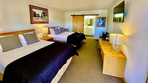 A hotel room with two beds, a desk, a chair, a TV, and a bathroom visible through a sliding door. - Old Montana Inn (Deer Lodge)