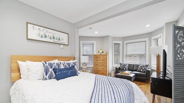 A modern bedroom with a large bed, a dresser, a TV, and a window with blinds. - North End Studio (Boston)