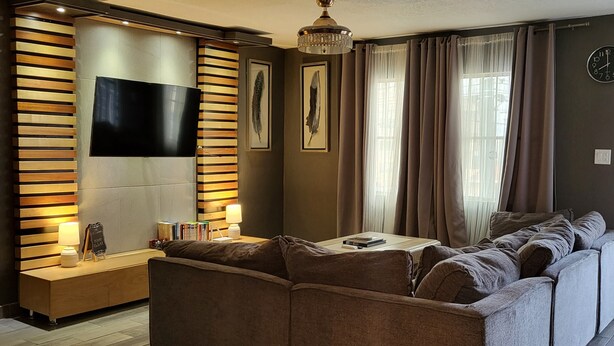 Ein modernes Wohnzimmer mit einem Ecksofa, einem Flachbildfernseher an einer Wand aus Holzpaneelen, einer Chandelieber und einem Bücherregal mit Büchern.