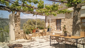Edificio de piedra con un patio que incluye una mesa, sillas, plantas en macetas y una pérgola cubierta de hiedra.