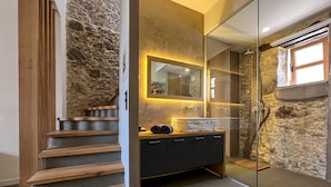 Un baño moderno con pared de piedra, escalera de madera, cabina de ducha de vidrio y tocador con espejo.