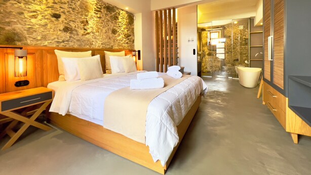 Una habitación de hotel moderna con una cama grande, cabecera de madera, pared de piedra y un baño visible a través de una puerta abierta.