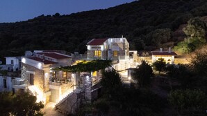 Una propiedad en una ladera con varias construcciones, incluyendo una casa principal con techo rojo y un edificio más pequeño con fachada amarilla. Hay muros de piedra y una escalera que conduce a la casa principal.