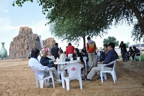 A group of people seated at tables outdoors with a large tree overhead, a rocky formation in the background, and a clear sky.