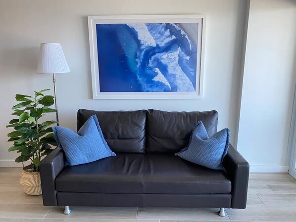 A dark leather sofa with two blue pillows, a framed abstract painting above it, and a potted plant to the left. - Luxury Manly Beachfront Apartment 3bdrs/2 bathrooms (Manly)