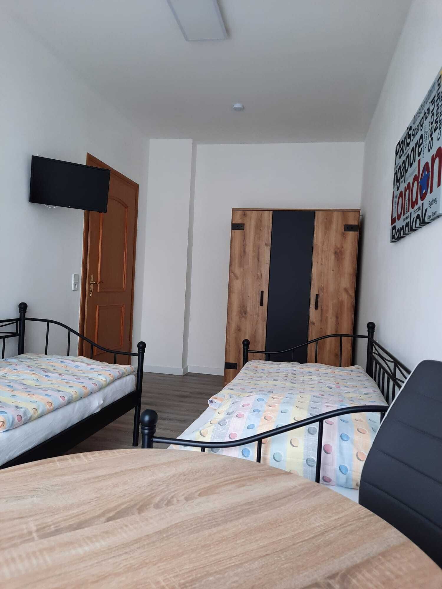 A room with two beds, a wooden table, a TV mounted on the wall, and a door.
