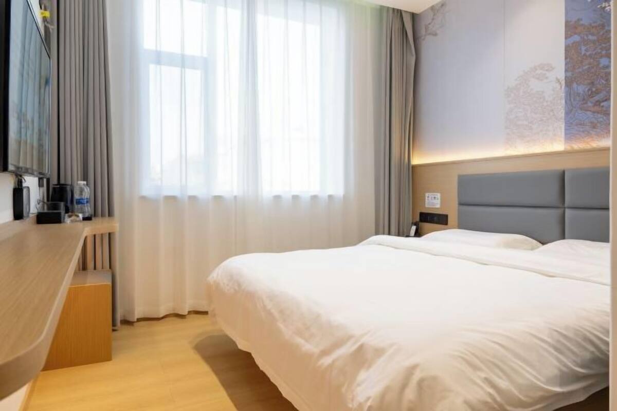 Une chambre d’hôtel comprenant un lit, un bureau, une chaise, une fenêtre avec des rideaux et un mur orné d’une fresque représentant un arbre.