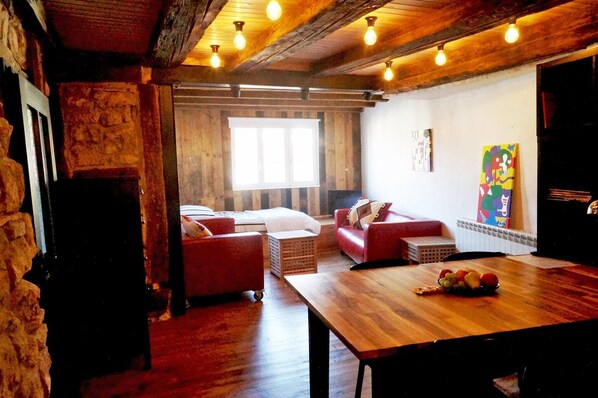 A cozy room with a wooden ceiling, a dining table with fruit, a red sofa, and a colorful abstract painting on the wall.