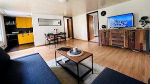 Una sala de estar moderna con sofá, mesa de centro y televisor. Hay un comedor con sillas y una mesa, y una cocina con armarios amarillos.
