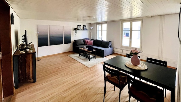 Un espacio habitable amplio con un sofá de color oscuro, una mesa de centro y una mesa de comedor con sillas. Hay ventanas grandes que dejan entrar luz natural y ofrecen vistas al exterior.