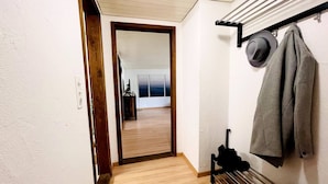 Un pasillo con piso de madera, un perchero con sombreros y una chaqueta, y una puerta que da a otra habitación.